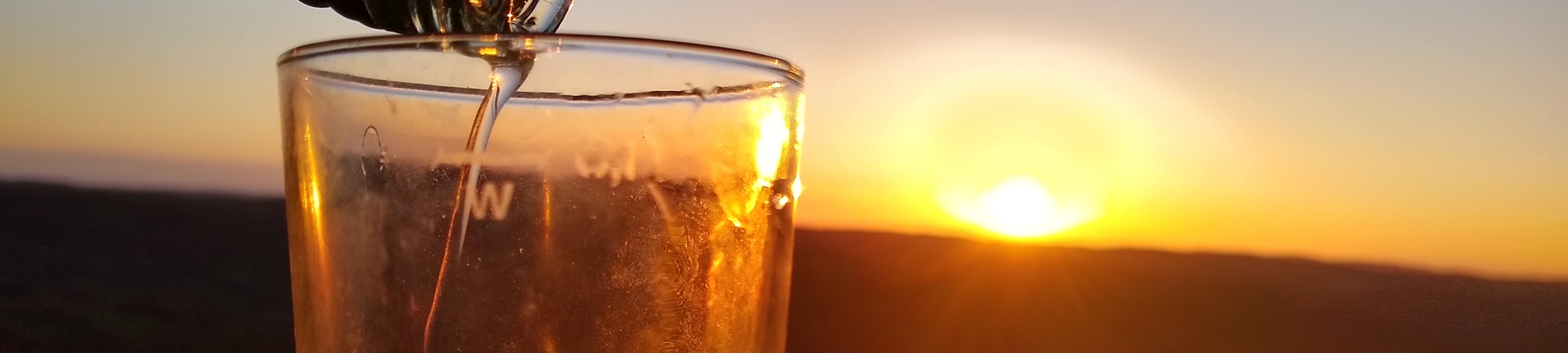 Flüssigkeit wird aus einer Flasche in ein Glas gegossen vor einem Sonnenuntergang über einer Landschaft